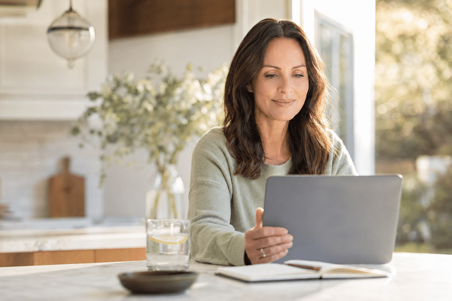 Woman in a gray sweater works on a silver laptop at a bright kitchen table, with a glass of water and an open notebook nearby.